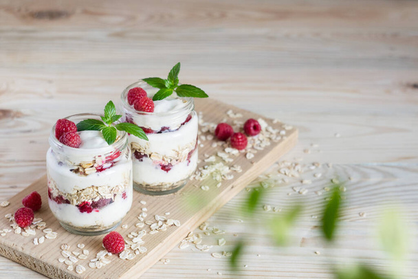 Two portions of homemade natural yogurt with oats and fresh raspberries in glass jars on a light wooden background. Concept of healthy breakfast. Horizontal orientation, selective focus, copyspace. - Фото, зображення