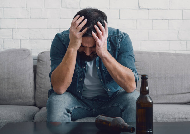 Sad alcoholic holds his head with hands, empty bottles on table - Photo, Image