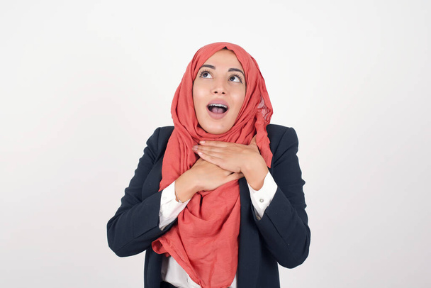 Счастливая красивая мусульманка носит черный блейзер, а хиджаб, улыбаясь изолированно на сером студийном фоне, имеет руки на груди возле сердца. Молодая эмоциональная женщина. Человеческие эмоции, концепция выражения лица. Вид спереди. - Фото, изображение