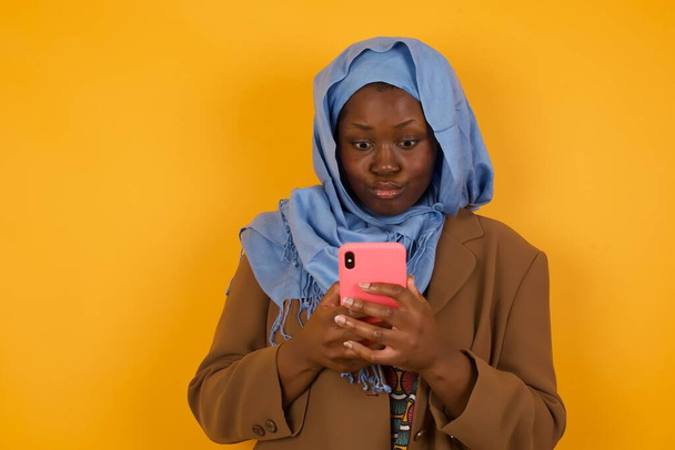 Vaaka laukaus melko nuori Afrikkalainen Amerikan musliminainen näyttää salakuunnellun silmät, pitää moderni älypuhelin, reseptejä unexpcted viestin ystävä, lukee muistutus, eristetty keltaisella taustalla. - Valokuva, kuva
