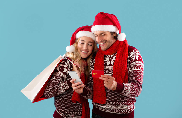 Loving young couple in winter outfits doing online Christmas shopping with credit card and smartphone on blue background - Foto, Imagen