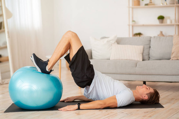 Athletic guy in sportswear and fitness tracker doing abdominal exercises in living room interior - Photo, Image
