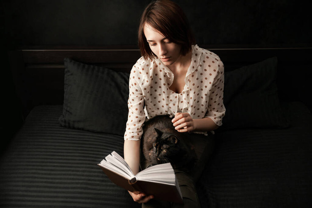 Mulher bonita lê um livro enquanto se senta na cama com um gato em suas pernas. Menina estudante estudando em casa - Foto, Imagem