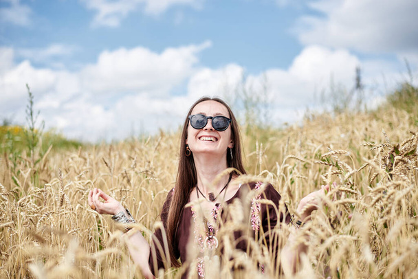 Young brunette hippie woman, wearing boho style clothes, sitting in the middle of wheat field with blue sky, meditating, thinking, relaxing, enjoying. Eco tourism concept. Nature protection. - Фото, зображення