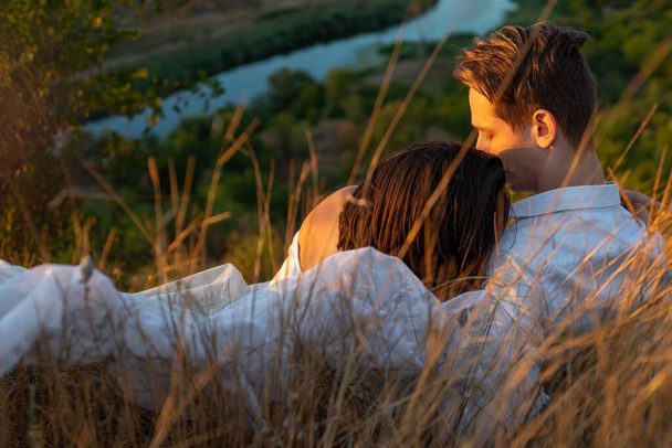草の上に寝そべっている男と女の美しいカップルの半分の肖像画。夏の夜の夕日を背景に。花嫁の白いドレス。草の分野で20歳の若いカップル。緑の草の上の男と,  - 写真・画像