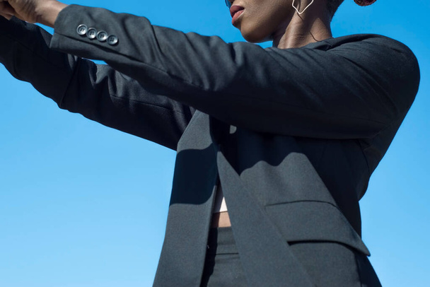 Joven mujer de negocios africana a la moda en una chaqueta de traje negro de negocios y pantalones posando mostrando detalles de un traje. En el fondo del cielo azul. Estilo de moda de negocios en un traje negro, en una chica negra. Estilo de negocio chica africana - Foto, imagen
