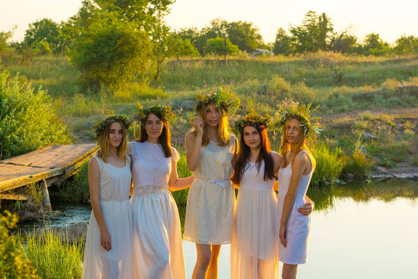 grupa młodych dziewcząt w wieku dwudziestu lat, w białych sukienkach, z wieńcami kwiatów, przytulających się, stojących przy zachodzie słońca nad stawem. Święto narodowe panny młodej, wieńce na głowie kwiatów, młode dziewczyny, koncepcja dziewictwa, wieczór panieński, I - Zdjęcie, obraz
