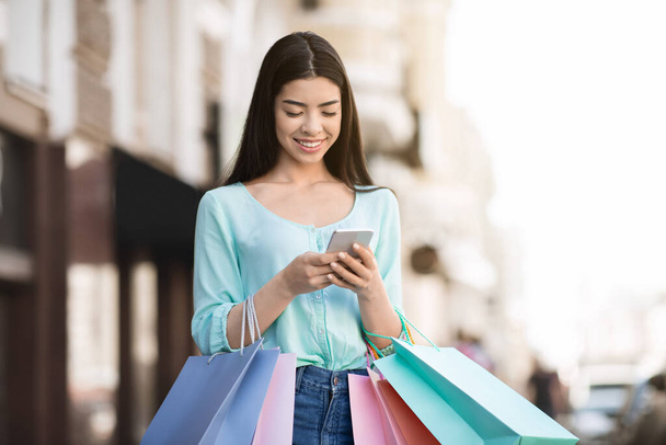 Joven Shopaholic chica asiática usando Smartphone al aire libre después de compras exitosas en el centro comercial - Foto, imagen