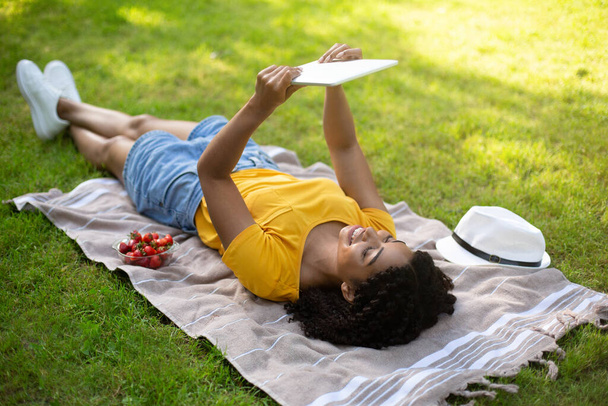 Funky African American girl with tablet watching educational video while lying on blanket at park - Photo, Image