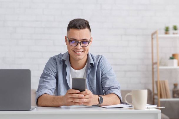 Wpisz telefon i pracuj online. Uśmiechnięty facet w koszuli patrzy na smartfona, siedzi przy stole z laptopem - Zdjęcie, obraz