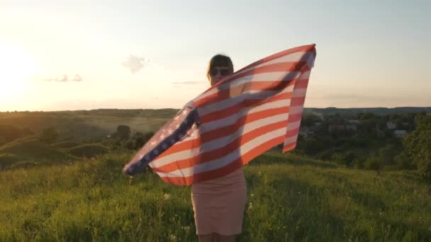 Jovem posando com bandeira nacional dos EUA ao ar livre ao pôr do sol. Menina positiva celebrando o dia da independência dos Estados Unidos. - Filmagem, Vídeo