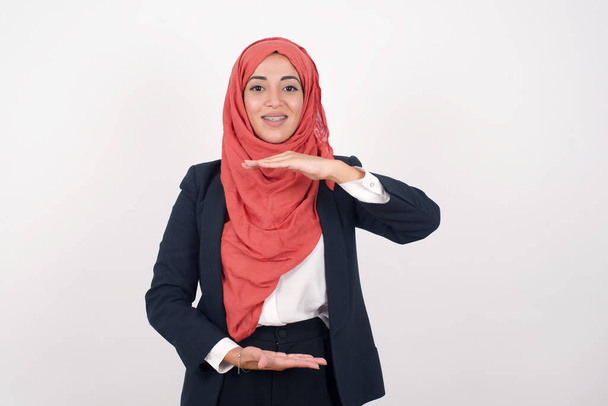 美しいイスラム教徒の女性は,黒いブレザーとヒジャーブーバーを身に着け,大きなサイズのサインを示し,シンボルを測定する手で身に着けています. カメラを見ている笑顔. 測定コンセプト. - 写真・画像