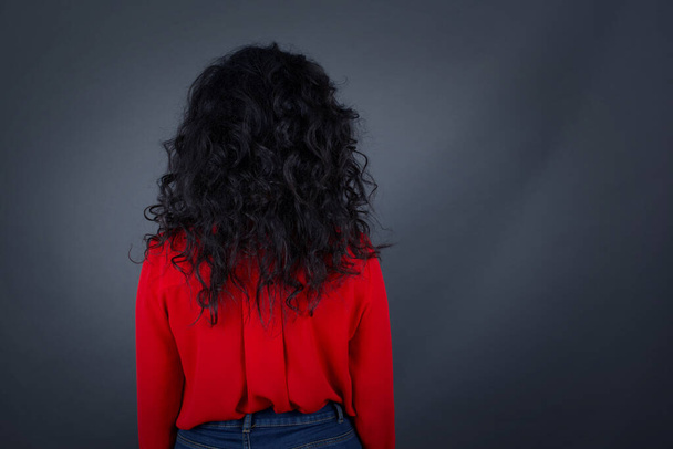 back view of brunette woman with curly hair wearing red shirt standing against gray wall. Studio Shoot. - 写真・画像
