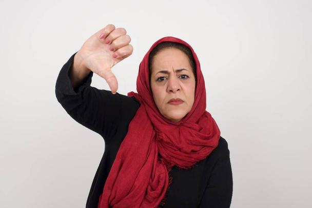 Discontent woman shows disapproval sign, keeps thumb down, expresses dislike, frowns face in discontent, dressed in white shirt, isolated over gray background. Body language concept. Middle-aged Arab woman wearing head scarf posing in studio - Photo, Image