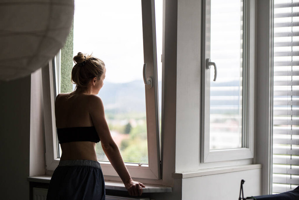 Home quarantine and social distancing during covid pandemic. Silhouette of lonley caucasian woman standing by window, anxiously looking out. Coronavirus infection, pandemics, disease outbreaks - Foto, Imagem