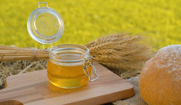 Close up honey in a glass jar. Traditional baked bread and sheaf of ripe rye ears on a fresh hay. Healthy food concept. Summer still life - Фото, зображення