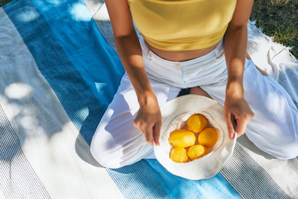 Top view of a young woman holds white headwear with lemons. Girl in white pants and yellow jersey holding a cotton basket of fresh-picked lemons sitting on the picnic blanket on a sunny day outdoors. - Foto, Bild