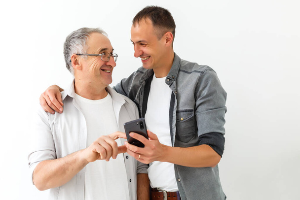 Happy two age generations men family old father embracing young grown adult son having fun enjoying using smart phone bonding watching funny social media video using mobile apps - Photo, Image