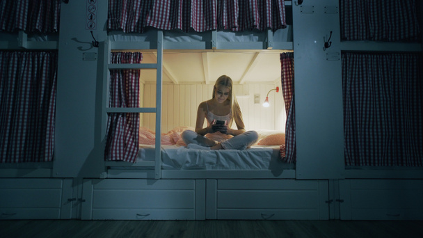 Young attractive woman sitting on the bed in the hostel room and using smartphone at night - Photo, Image