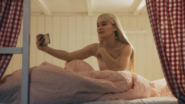 Közelkép fiatal vonzó nő ül az ágyon a hostel szobában, és használja okostelefon, amelynek video chat - Fotó, kép