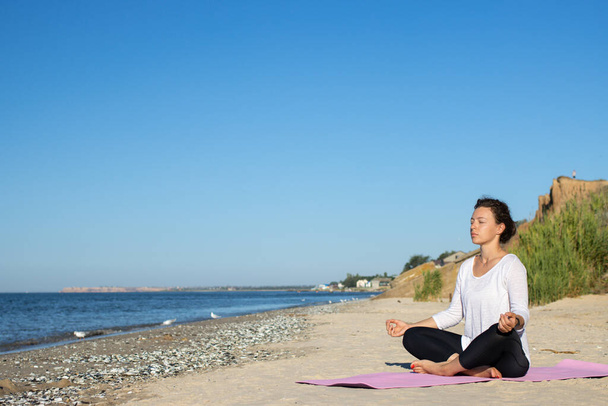 Young beautiful brunette woman practicing yoga, sitting alone in lotus position asana and meditating with closed eyes at seashore in the summer morning. Relax, wellness, healthy lifestyle. Copy space. - Foto, Imagem