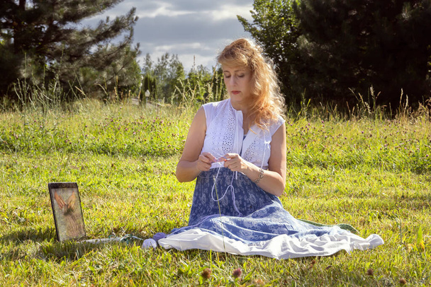 女性は、編み針の上に毛糸の服を編む公園で草の上に座って、レッスンを見たり、ラップトップを通して編み物を教えています。距離学習 - 写真・画像