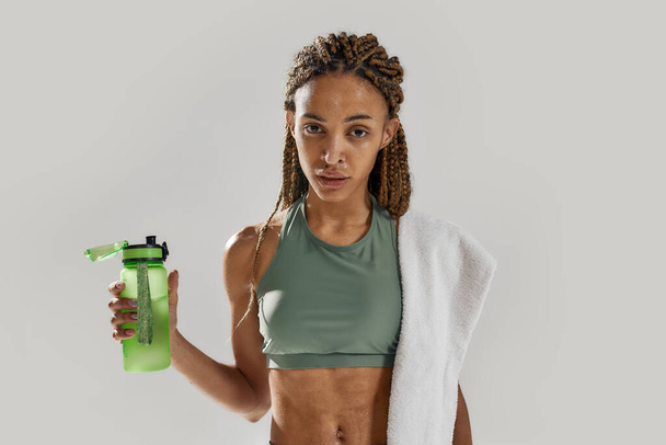 Young sportive mixed race woman looking at camera, holding protein shake cocktail bottle after intensive training isolated over grey background - Фото, зображення
