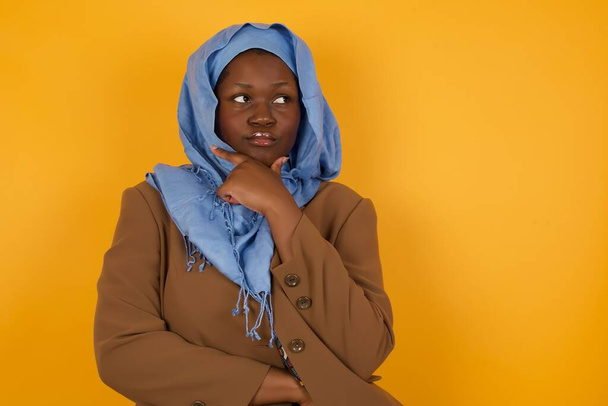 Απομονωμένο πορτρέτο της κομψής νεαρής Αφροαμερικανίδας μουσουλμάνα με το χέρι κάτω από το πηγούνι και κοιτάζοντας πλάγια με αμφίβολη και σκεπτικιστική έκφραση, ανησυχία και αμφιβολία. Στέκεται σε εσωτερικούς χώρους πάνω από κίτρινο φόντο. - Φωτογραφία, εικόνα