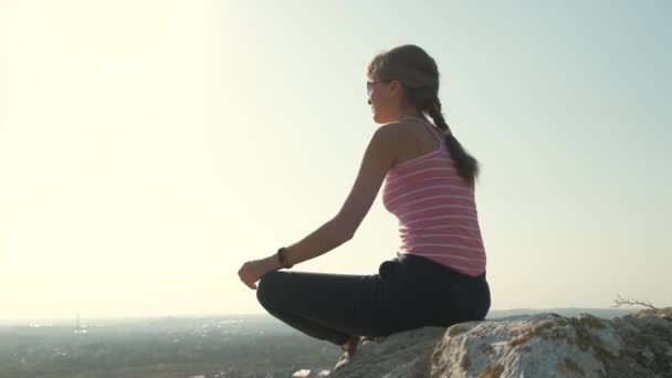 Молодая расслабленная женщина сидит на открытом воздухе на большом камне наслаждаясь теплым летним днем. Девушка медитирует и отдыхает на природе на закате. - Кадры, видео