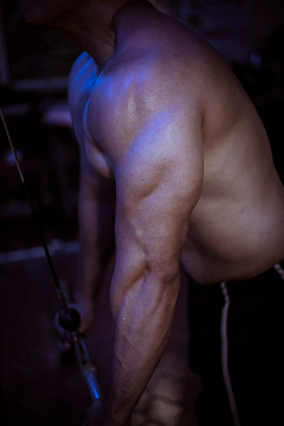 Um homem anônimo em topless treina seu tríceps com empurrões de máquina. Treinamento intenso. Configuração escura com iluminação azul. Corpo suado após meia hora de exercício. - Foto, Imagem