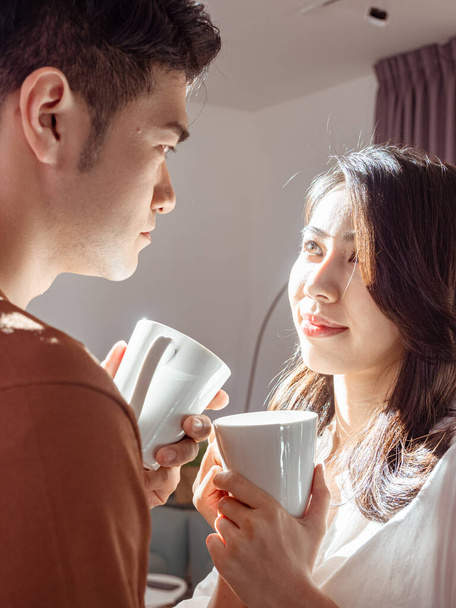 Νεαρή Ασιάτισσα και άντρας απολαμβάνουν να περνούν χρόνο μαζί στο σπίτι με ένα φλιτζάνι καφέ στα χέρια. - Φωτογραφία, εικόνα