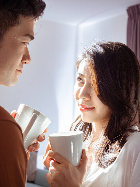 Young asian woman and man are enjoying spending time together at home with cup of coffee in hands. - Photo, Image