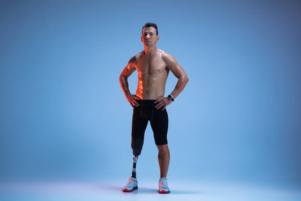 Athlete with disabilities or amputee isolated on blue studio background. Professional male sportsman with leg prosthesis training with weights in neon - Zdjęcie, obraz