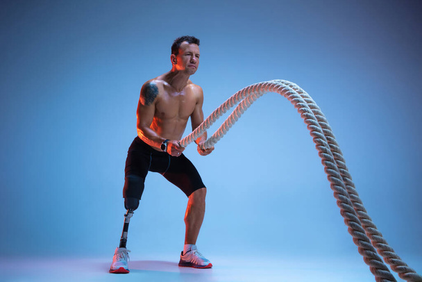 Athlete with disabilities or amputee isolated on blue studio background. Professional male sportsman with leg prosthesis training with weights in neon - Foto, imagen