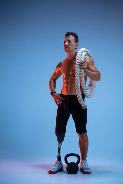 Athlete with disabilities or amputee isolated on blue studio background. Professional male sportsman with leg prosthesis training with weights in neon - Photo, image
