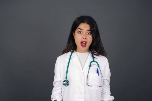 Emotional attractive  doctor  with opened mouth expresses great surprisment and frighteness, poses against white concrete background, stares at camera. Unexpected shocking news and human reaction. - Φωτογραφία, εικόνα
