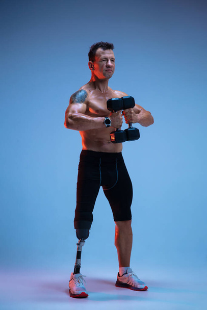 Athlete with disabilities or amputee isolated on blue studio background. Professional male sportsman with leg prosthesis training with weights in neon - Fotografie, Obrázek