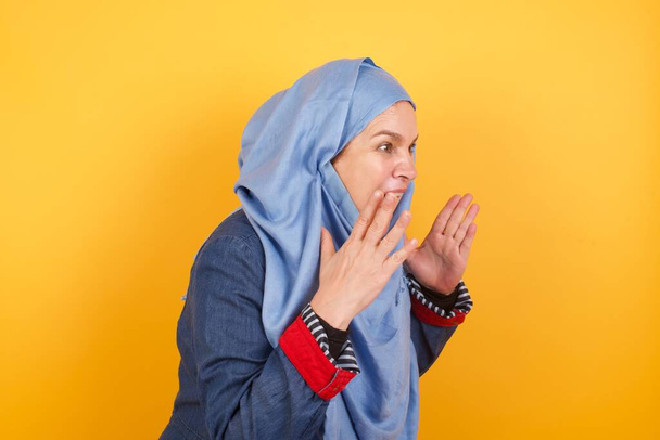 Porträt einer muslimischen Frau mittleren Alters im Hijab, die vor gelbem Hintergrund steht, mit schockiertem Gesichtsausdruck, die Hände vor dem Gesicht hält, schreit und seitwärts auf etwas Erstaunliches blickt. - Foto, Bild