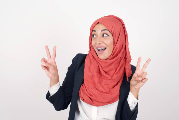 Isolated shot of Beautiful muslim woman  wears black blazer and hijab makes peace or victory sign with both hands, dressed in casual clothes, feels cool has toothy smile, isolated over gray background. People and body language. - Foto, afbeelding
