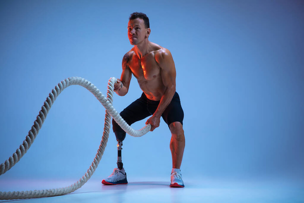 Athlete with disabilities or amputee isolated on blue studio background. Professional male sportsman with leg prosthesis training with weights in neon - Foto, Bild