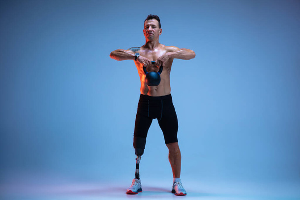 Athlete with disabilities or amputee isolated on blue studio background. Professional male sportsman with leg prosthesis training with weights in neon - Foto, Imagem