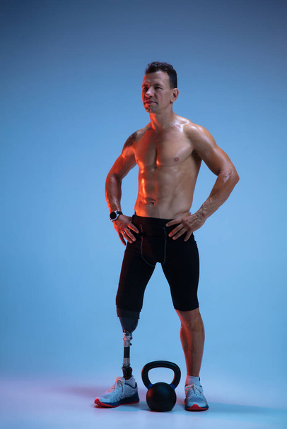 Athlete with disabilities or amputee isolated on blue studio background. Professional male sportsman with leg prosthesis training with weights in neon - Foto, afbeelding