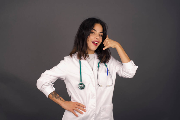 Aantrekkelijke jonge arts vrouw in casual kleding imiteert telefoongesprek, houdt de hand dicht bij het oor alsof het houden van mobiele telefoon, heeft zelfverzekerde gezichtsuitdrukking, geïsoleerde - Foto, afbeelding
