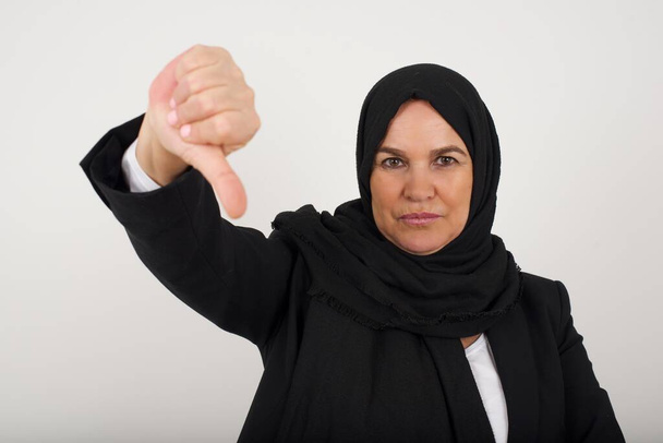 Discontent muslim woman in black hijab shows disapproval sign, keeps thumb down, expresses dislike, frowns face in discontent, dressed in white shirt, isolated over gray background. Body language concept. - Photo, Image