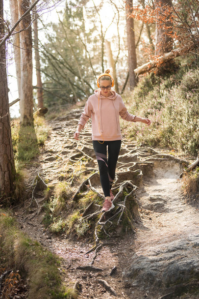 Active sporty woman running in autumn fall forest jumping over the roots on the path. Healthy lifestyle image of young active caucasian woman jogging outside in nature - Photo, Image