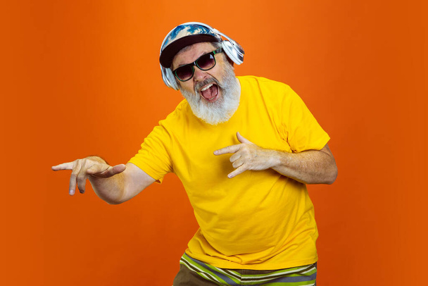 Starszy hipster korzystający z urządzeń, gadżety na pomarańczowym tle. Techniczna i radosna koncepcja stylu życia osób starszych - Zdjęcie, obraz