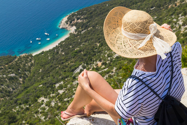 Aktivní sportovní žena na letních prázdninách sedící na staré kamenné zdi ve vesnici Lubenice, se slaměným kloboukem a plážovým batohem s krásným výhledem na pobřeží ostrova Cres, Chorvatsko - Fotografie, Obrázek