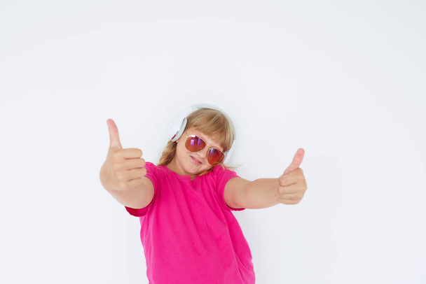 blonde teenage girl in headphones, sunglasses and a bright pink T-shirt stretched out her hands with thumbs up against a white wall - Foto, afbeelding