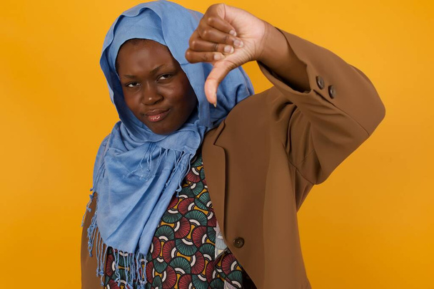 Discontent African American muslim woman shows disapproval sign, keeps thumb down, expresses dislike, frowns face in discontent, dressed in white shirt, isolated over yellow background. Body language concept. - Photo, Image