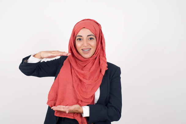 Beautiful muslim woman  wears black blazer and hijab over isolated background gesturing with hands showing big and large size sign, measure symbol. Smiling looking at the camera. Measuring concept. - Zdjęcie, obraz